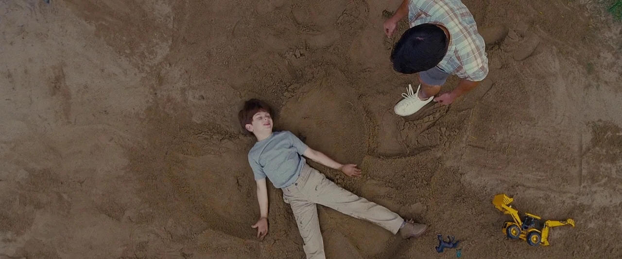 Stoker (2013) movie still: wide — A flat view of a sand pit with two individuals present. A child is lying on his back while an adult stands over…