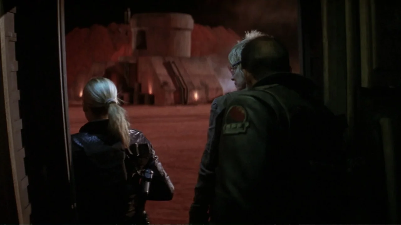 Ghosts of Mars (2001) movie still: medium — A dark, interior threshold overlooks a desert compound lit by deep red glowing lights. A woman and a man…