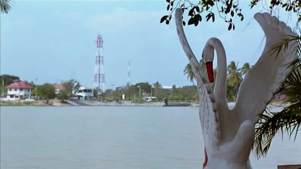 Tropical Malady (2004) movie still: wide — A calm lake sits in front of a distant shore with houses and towers. A large white statue of a swan…