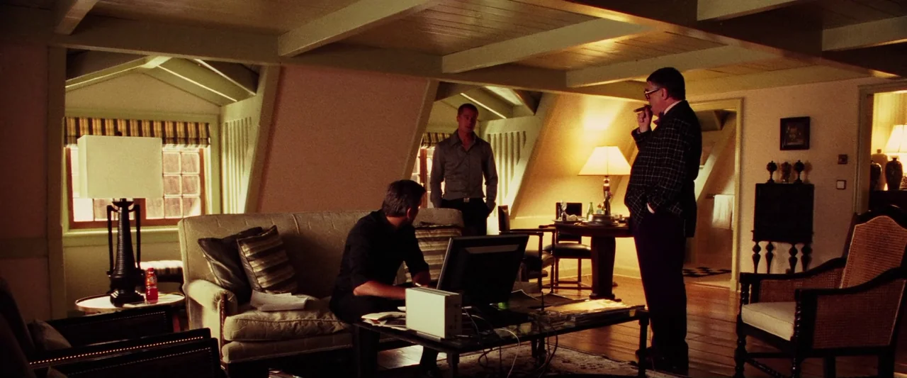 Ocean’s Twelve (2004) movie still: wide — A hotel suite features beige upholstered furniture, wood-beamed ceilings, and multiple lamps. Three men…