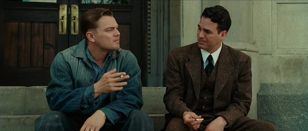 Shutter Island (2010) movie still: medium — Two men sit together on concrete steps in front of a heavy wooden door. They wear mid-20th century…