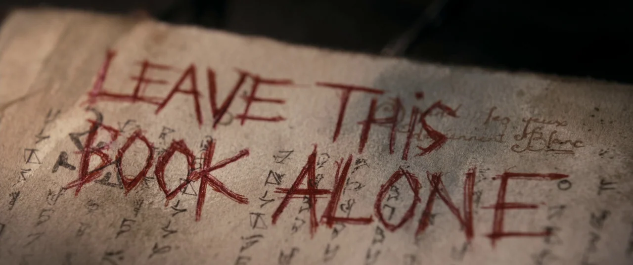 Evil Dead (2013) movie still: closeup — An ancient parchment page features handwritten symbols and deep red ink text. The words Leave This Book…