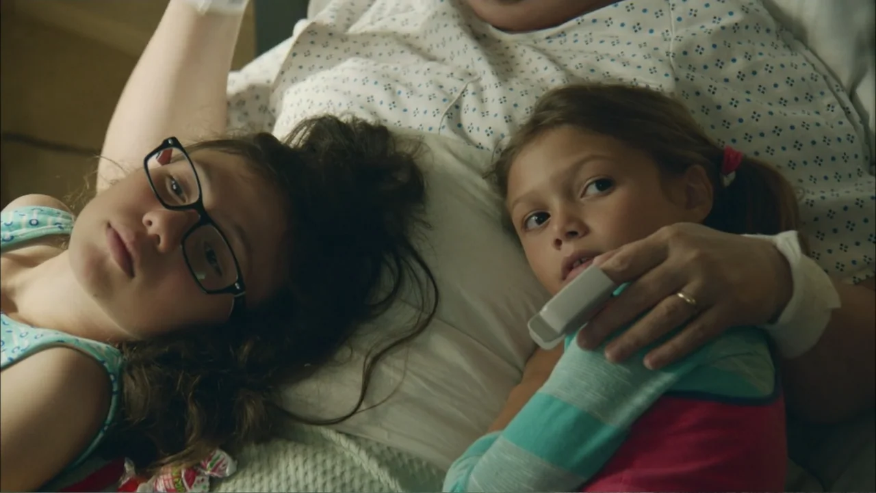 The Hollars (2016) movie still: closeup — A hospital bed with white patterned sheets holds two young children lying down. One child wears glasses…