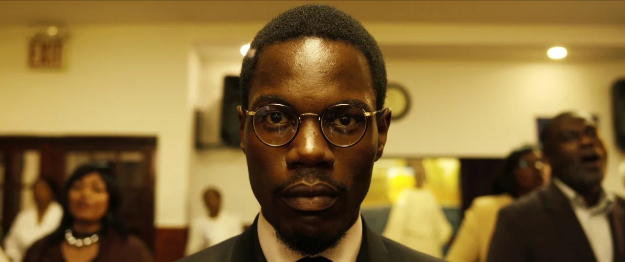 Da Sweet Blood of Jesus (2014) movie still: closeup — A man stands in a dimly lit, nondescript room with blurred people in the background. He is…