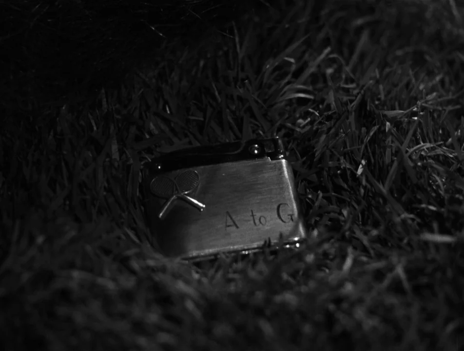 Strangers on a Train (1951) movie still: closeup — A silver lighter featuring a tennis racket emblem sits nestled within thick, dark grass. The…