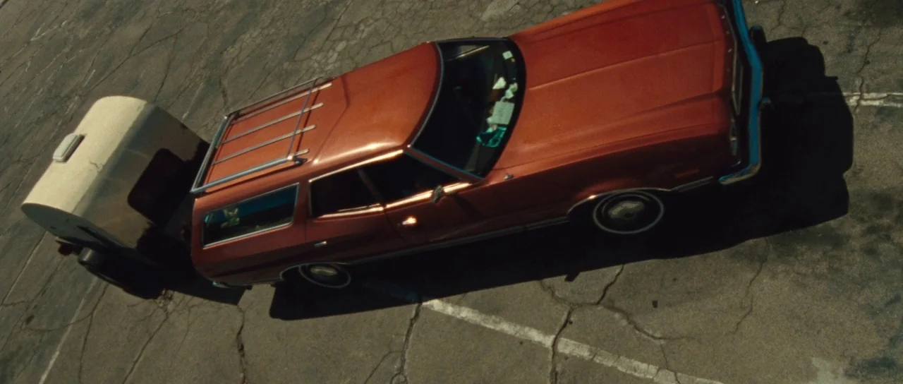 The Runaways (2010) movie still: wide — A vintage orange station wagon with a small, white, rounded trailer parked on asphalt. Large cracks run…