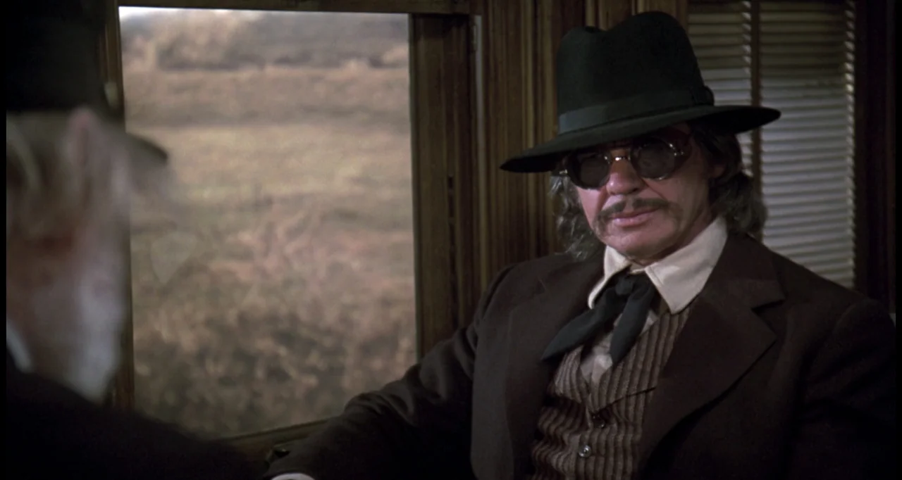 The White Buffalo (1977) movie still: medium — A passenger train car interior features wood paneling and a large window looking outside. A man in a…