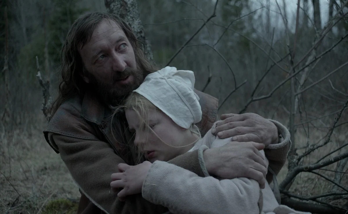 The Witch (2015) movie still: medium — A dense, leafless forest area with gray, muted tones and natural brush. A bearded man embraces a young girl…