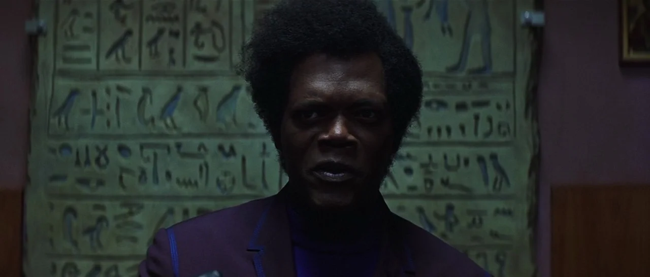 Unbreakable (2000) movie still: closeup — A man stands before a framed piece of papyrus decorated with Egyptian hieroglyphs. The subject gazes…