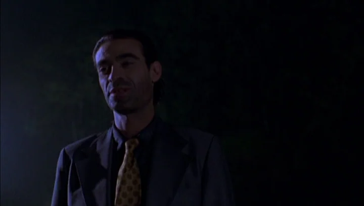 Highway 61 (1991) movie still: medium — A man stands in a dark, poorly lit exterior space at night. He wears a formal suit and looks ahead into the…