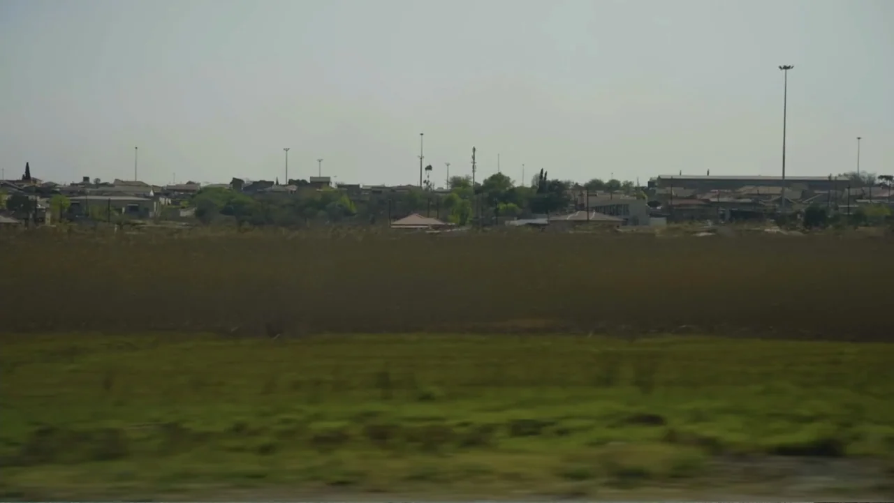 African Throne World Tour (2024) movie still: establishing — A flat field of dry grass sits in front of a sprawling suburban settlement. Numerous…