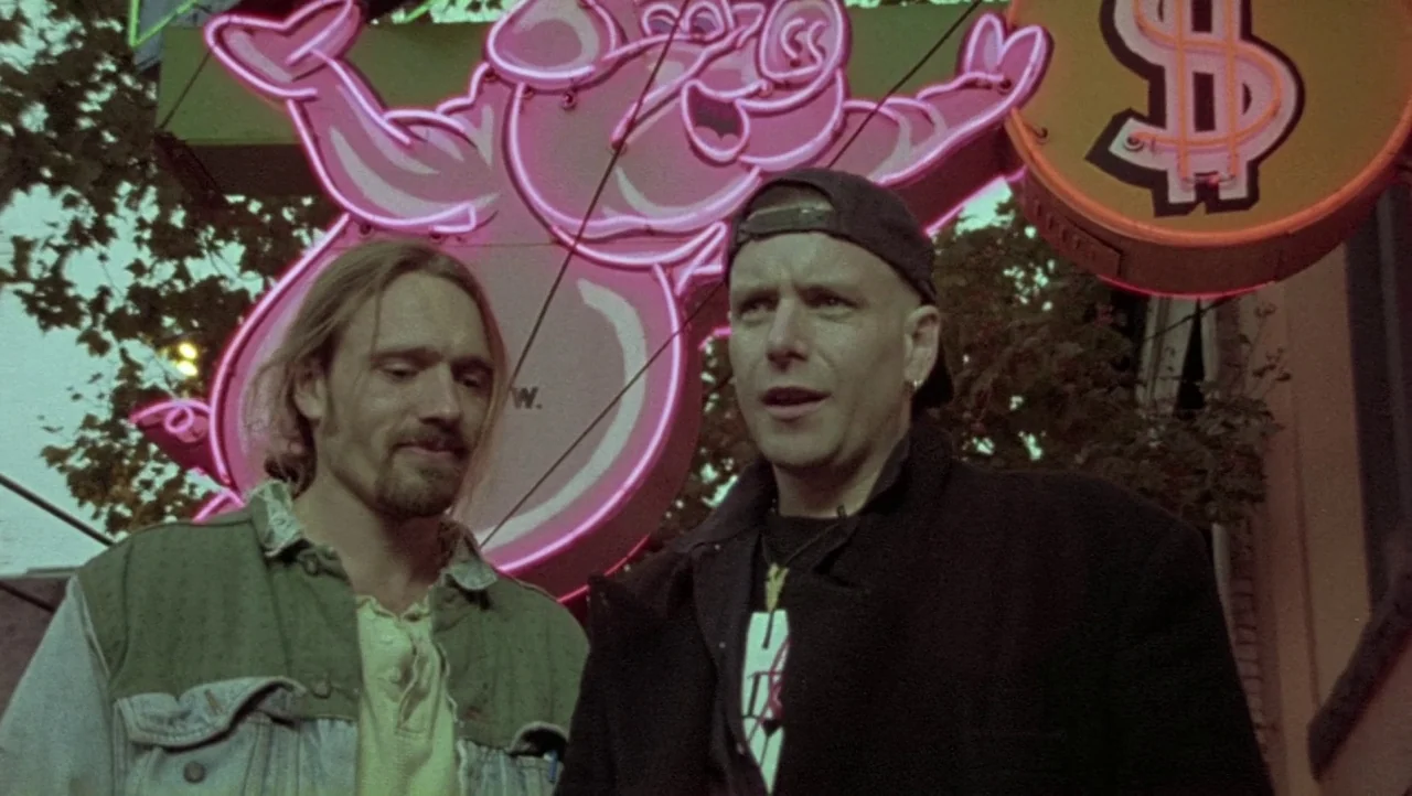 Hard Core Logo movie still: medium — An exterior night view featuring a bright pink pig neon sign and a dollar sign. Two men stand in the foreground…