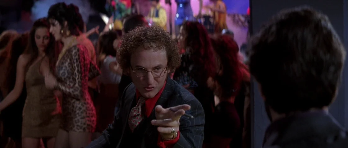 Carlito’s Way (1993) movie still: medium — A crowded, dimly lit nightclub interior with dancers visible in the blurry background. A man in a suit…