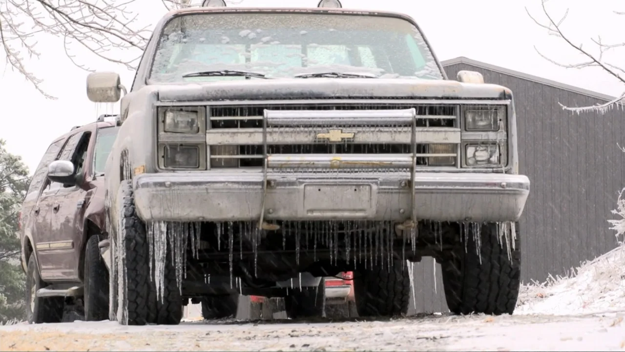 Meth Storm (2017) movie still: medium — A silver pickup truck and a dark maroon SUV sit parked in a snowy yard. Both vehicles are covered in thick…