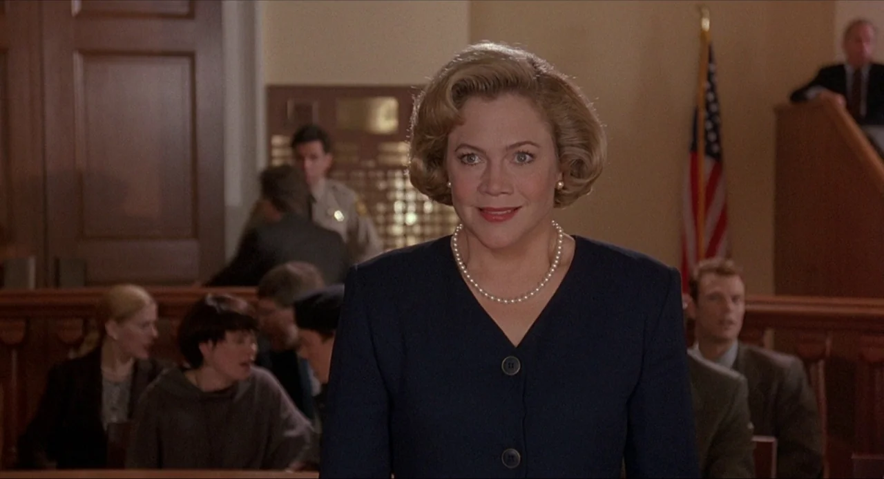Serial Mom (1994) movie still: medium — A courtroom interior with wooden barriers and an American flag in the background. A woman in a dark blazer…