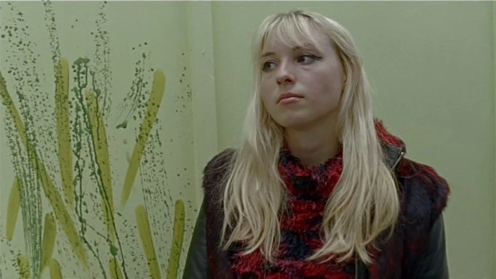Import Export (2007) movie still: medium — A light green wall features vertical paint drips and brush strokes in yellow and green. A woman with…