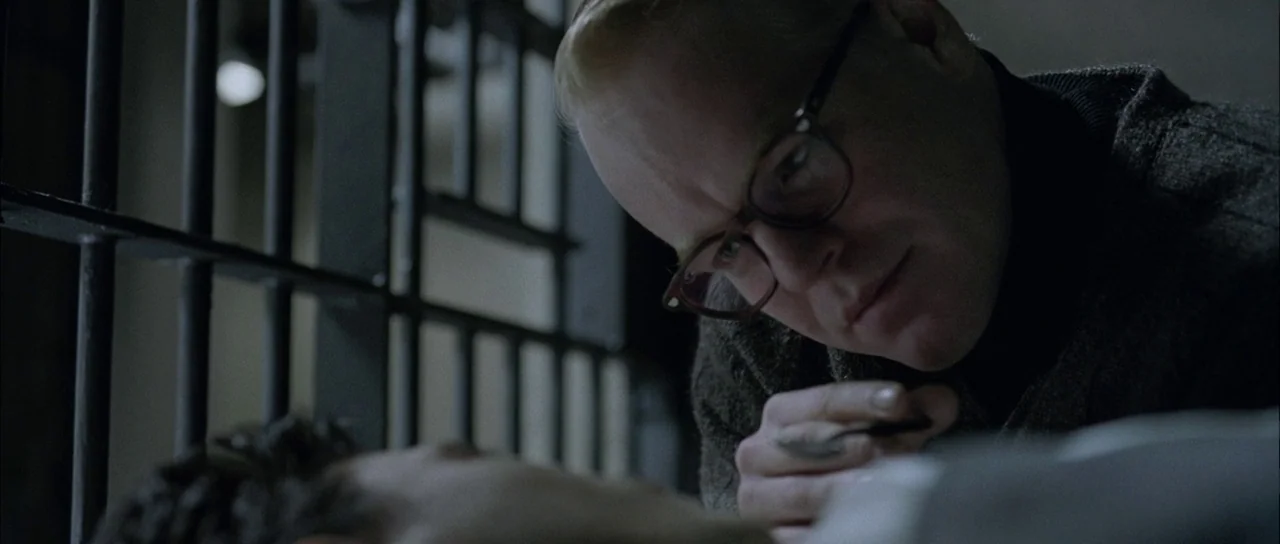 Capote (2005) movie still: closeup — A dimly lit room features heavy metal bars visible in the out-of-focus background. A man leans forward to…