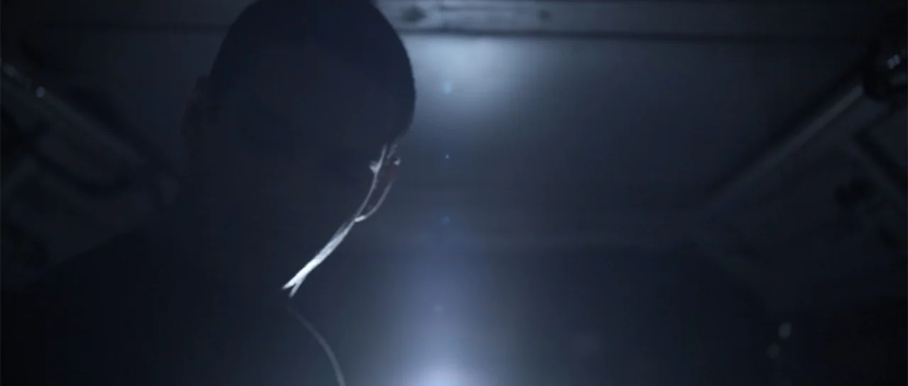 Astronaut: The Last Push (2012) movie still: closeup — A dark, dimly lit interior space with minimal architectural details visible. A person's…