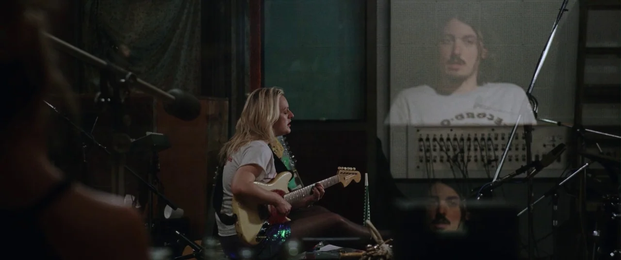 Her Smell (2018) movie still: medium — A dimly lit sound booth contains recording equipment and audio cables on the floor. A woman plays guitar…