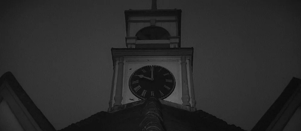 The Elephant Man (1980) movie still: establishing — A dark clock tower silhouette rises against a featureless night sky. The clock face shows Roman…
