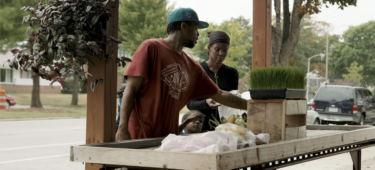 10 Billion: What's On Your Plate (2015) movie still: medium — A wooden produce stand sits on a suburban sidewalk during the day. A man serves fresh…