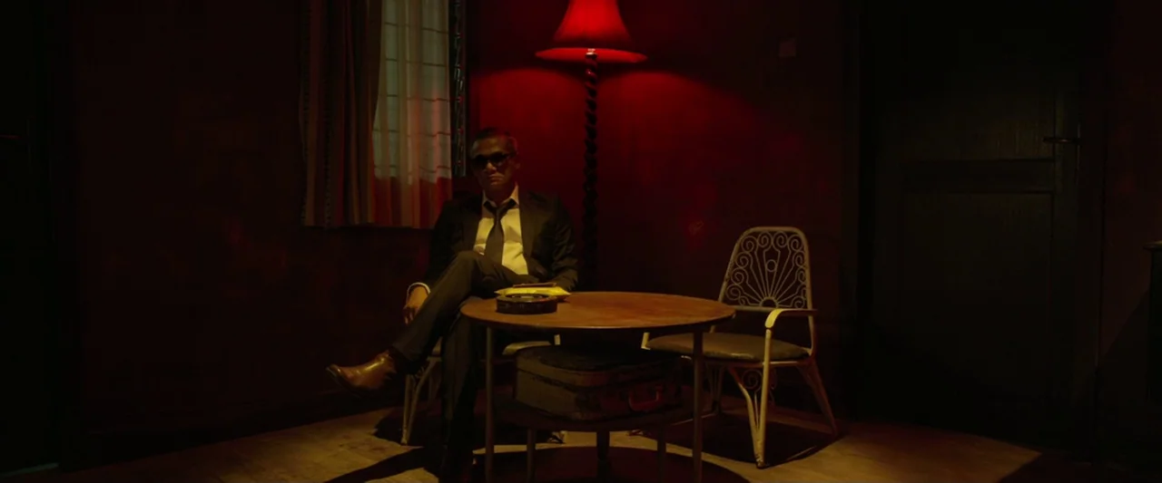 The Raid 2 (2014) movie still: medium — A dark, minimally furnished room is washed in intense red light from a lamp. A man in a dark suit sits at a…