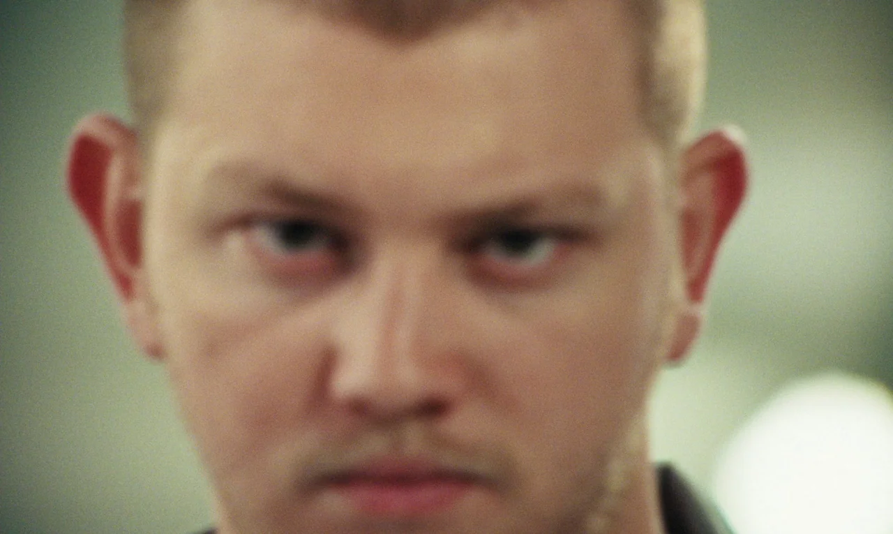 Maldoror (2024) movie still: closeup — An out-of-focus background obscures the details of an indoor setting. A man faces the camera with a direct…