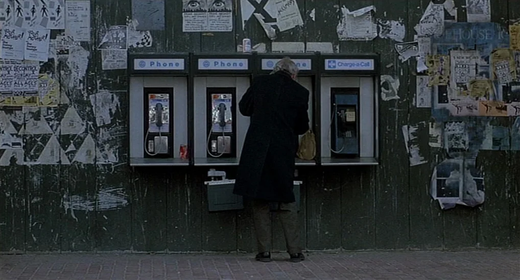 The Verdict (1982) movie still: fullbody — A vertical wooden wall is covered in weathered paper posters and multiple public payphones. An older man…
