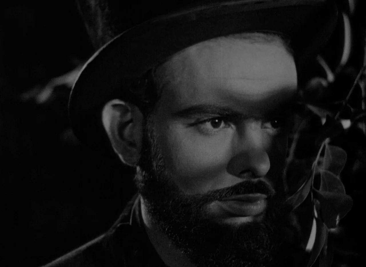 At Midnight I’ll Take Your Soul (1964) movie still: closeup — A man in a top hat stands among dark leaves at night. He looks off-screen while hard…