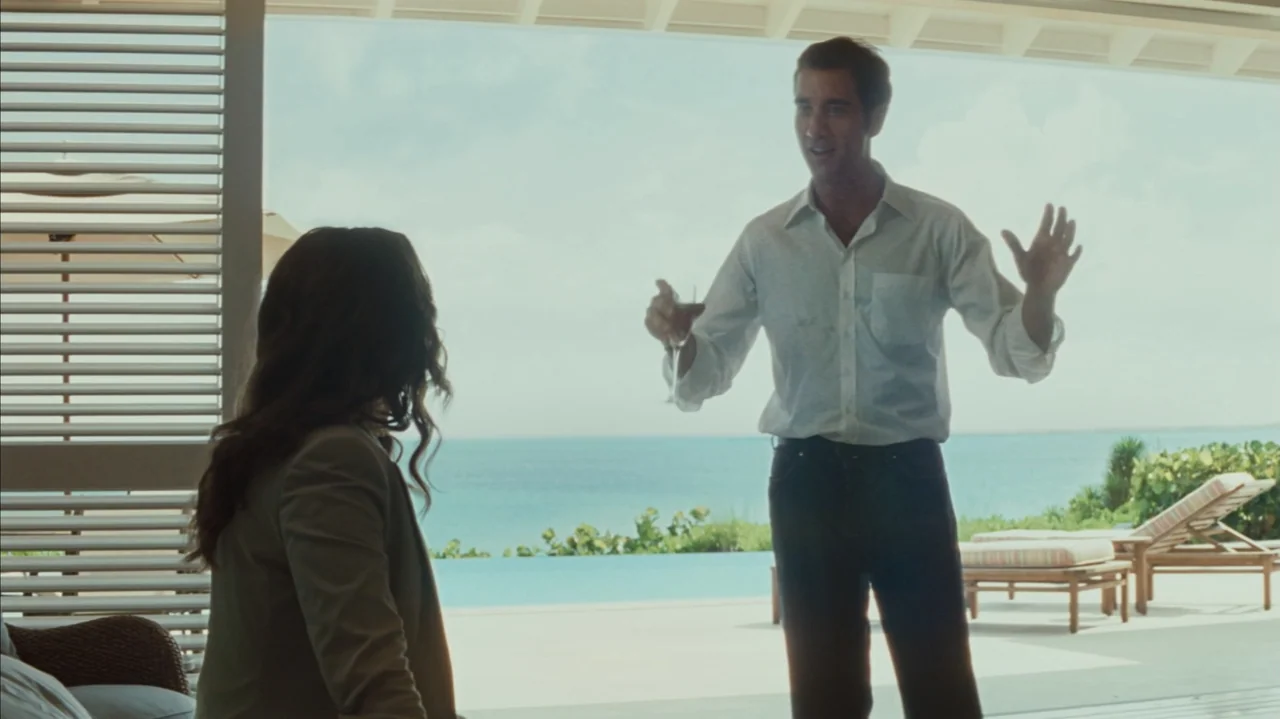 Duplicity (2009) movie still: fullbody — A bright patio opens toward an infinity pool overlooking a calm tropical sea. A man stands in a white shirt…