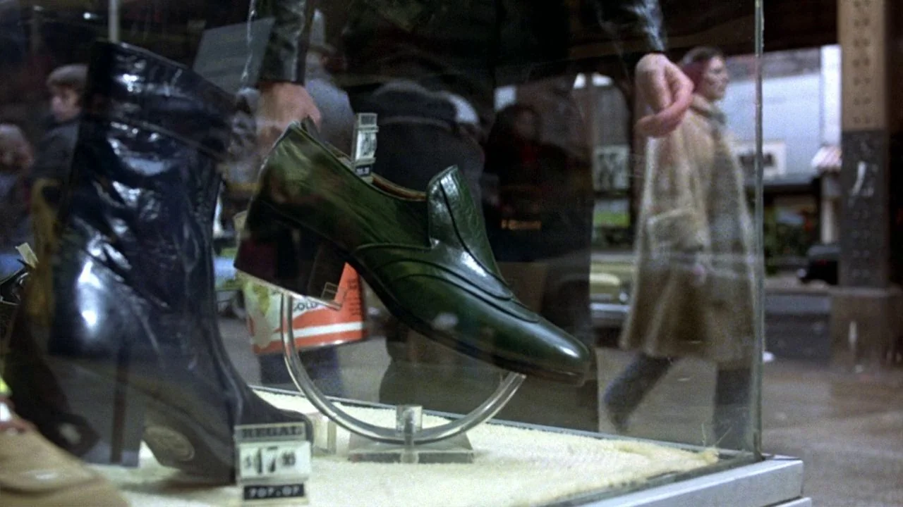 Saturday Night Fever (1977) movie still: closeup — A glass storefront display contains a green loafer on a clear plastic stand. Reflections on the…