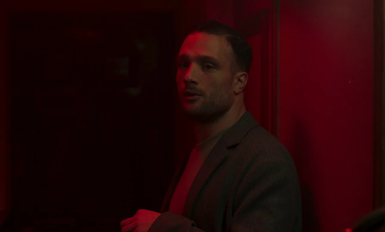 It Is In Us All (2022) movie still: medium — A man stands in a dark narrow corridor lit by an intense red light source. He wears a blazer over a…