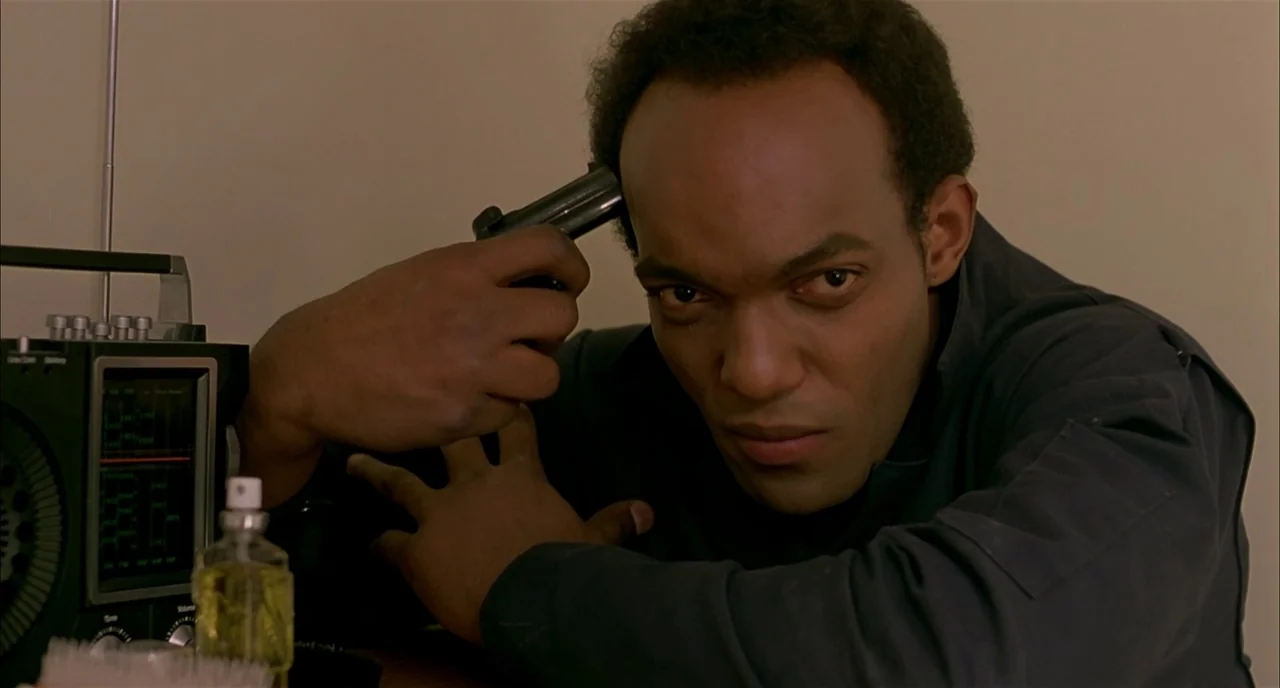 Dawn of the Dead (1978) movie still: medium — An interior space with a beige wall serving as a plain background. A man holds a small black handgun…