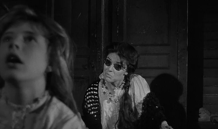 The Miracle Worker (1962) movie still: medium — A dimly lit room with dark wooden paneling in the background. A woman in dark glasses watches a…