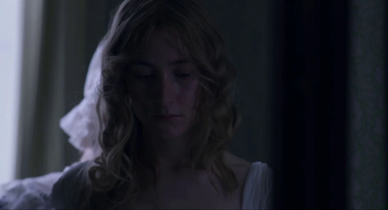 Ammonite (2020) movie still: closeup — A dimly lit room with soft light emanating from an off-screen window source. A young woman with…
