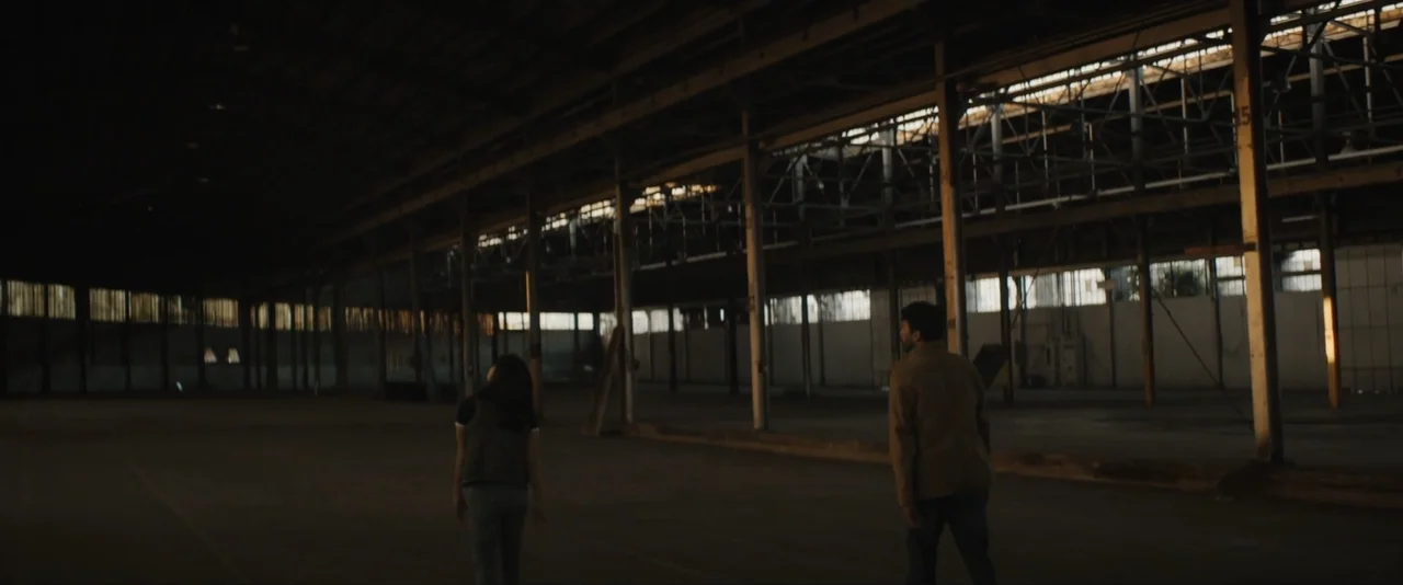 Stalker (2020) movie still: wide — An enormous, abandoned industrial warehouse with high ceilings and rows of metal support columns. Two individuals…