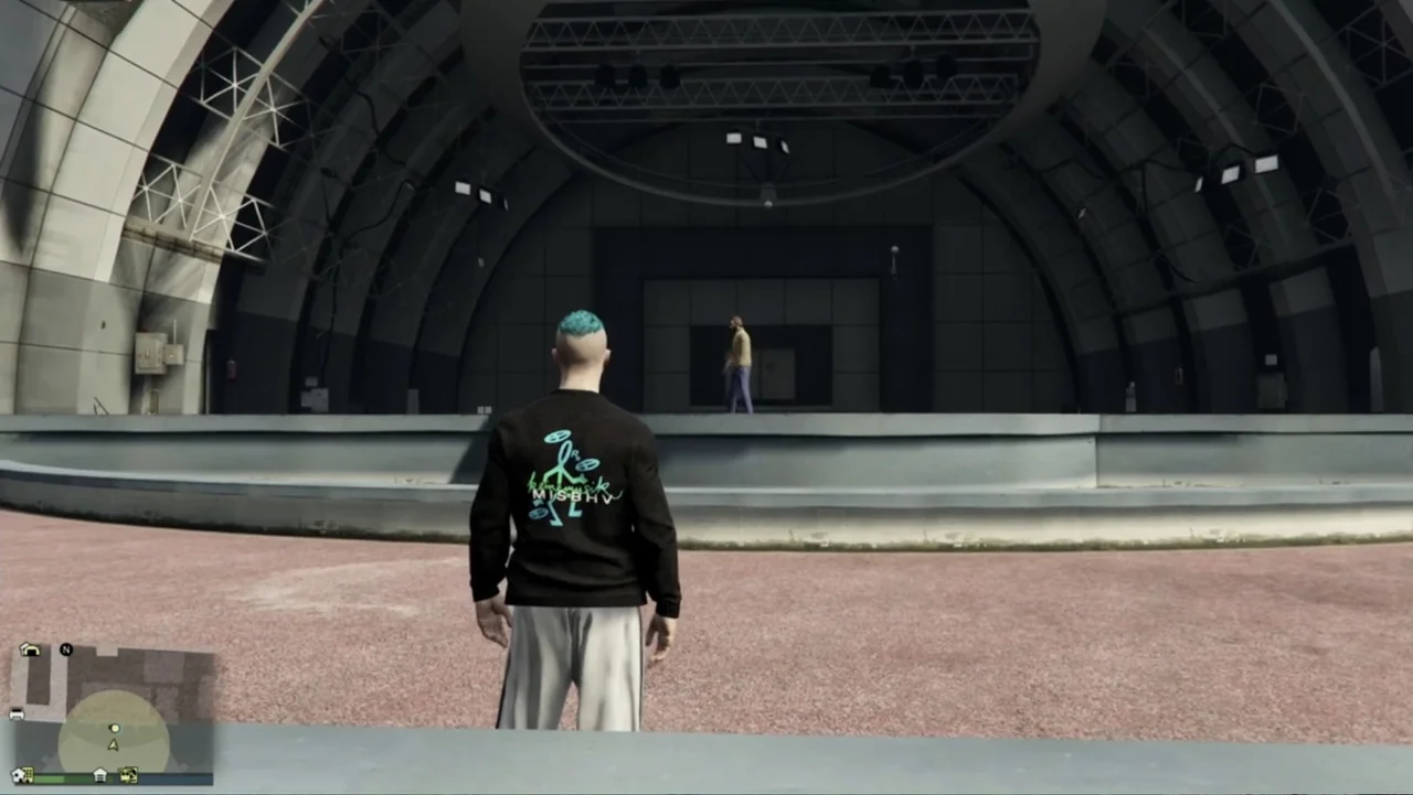 Grand Theft Hamlet (2024) movie still: medium — An outdoor amphitheater features a curved concrete stage area under a large archway. One person…