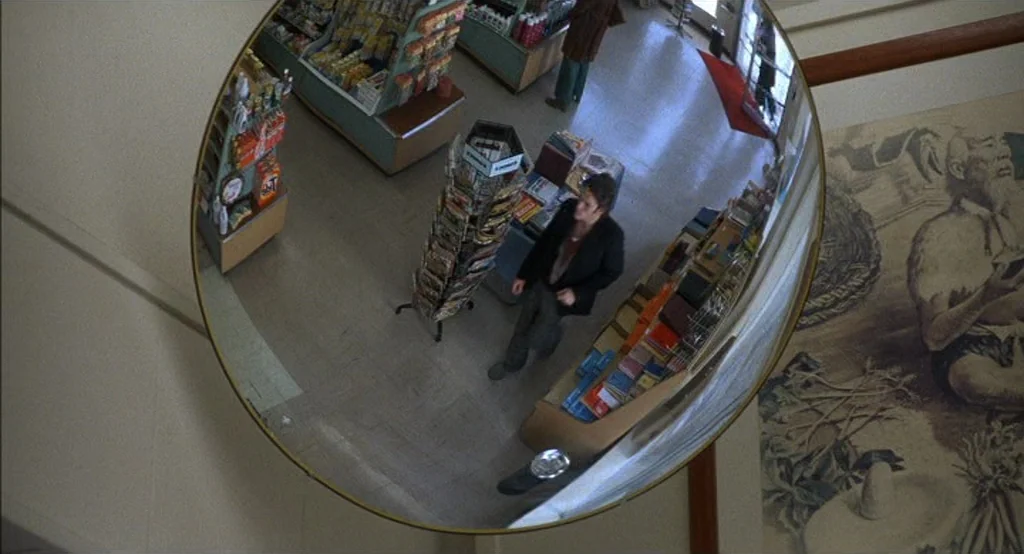 Drugstore Cowboy (1989) movie still: wide — A view of a retail space reflected in a circular security mirror. A man walks through the aisles past a…