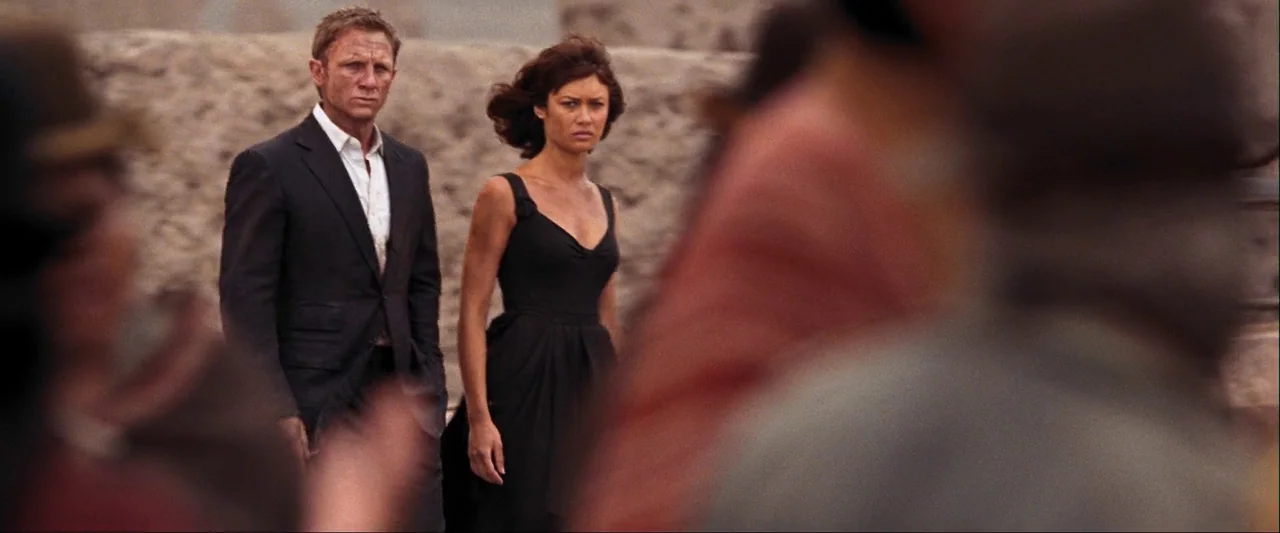 Quantum of Solace (2008) movie still: medium — A man and woman stand before a textured, arid-colored stone wall in a plaza. They watch events…