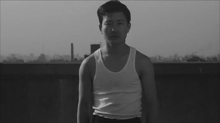 Death By Hanging (1968) movie still: medium — An urban rooftop with a low parapet wall against a blurred distant city skyline. A young man stands in…