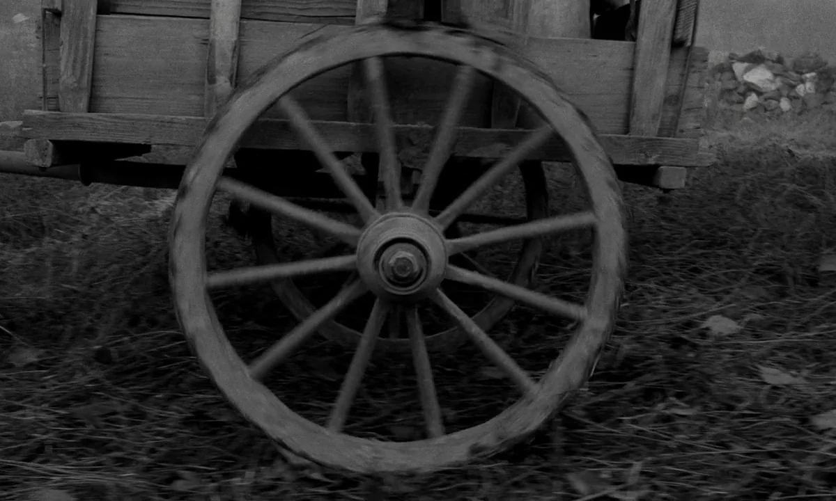 The Turin Horse (2011) movie still: closeup — A wooden cart wheel rolls across a patch of dry grass and dirt. The motion is captured in a close side…