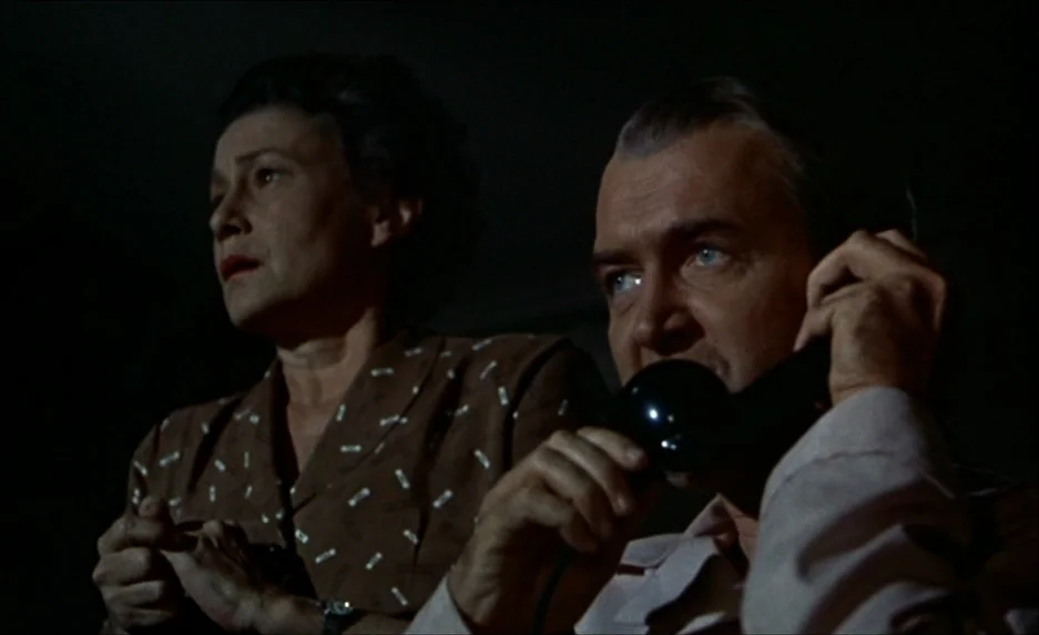 Rear Window (1954) movie still: medium — A dimly lit room at night with dark shadows obscuring the background. A man on the phone and a woman beside…