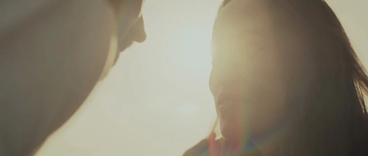 Meadowland (2015) movie still: closeup — Two individuals stand in an outdoor setting illuminated by bright, hazy golden sunlight. A woman is seen…