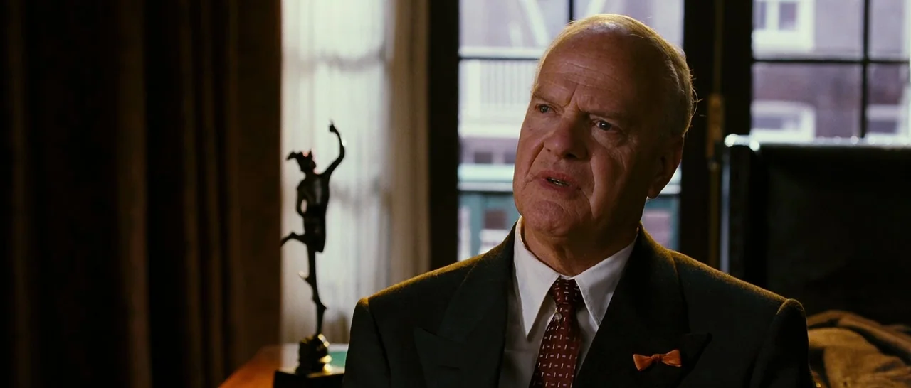 Black Book (2006) movie still: medium — An interior hotel suite features dark heavy curtains and a small bronze statue. An elderly man in a suit…