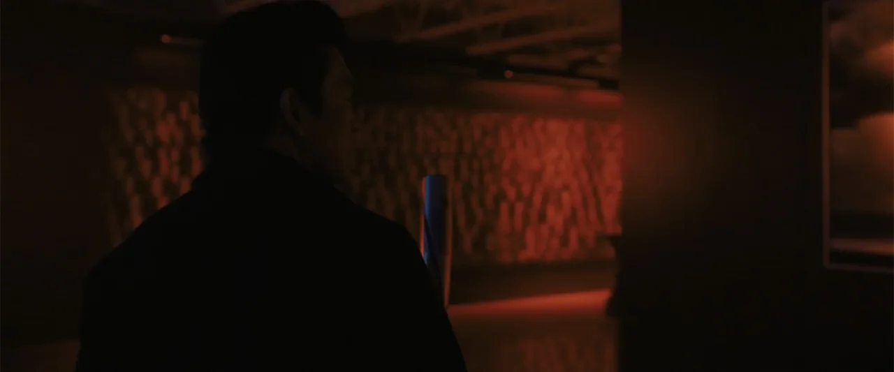 Afraid (2024) movie still: medium — A dark corridor features a wall illuminated by vibrant red neon light. A man wearing a dark jacket stands in the…