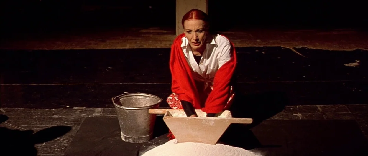 All About My Mother (1999) movie still: medium — A woman kneels on a dark floor in a minimalist performance space. She sifts white flour through a…