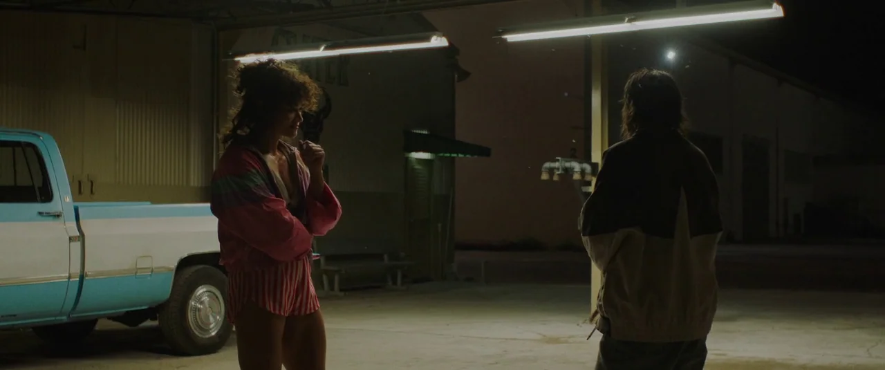 Love Lies Bleeding (2024) movie still: medium — A night setting at a service station under bright, industrial fluorescent overhead lights. A woman…