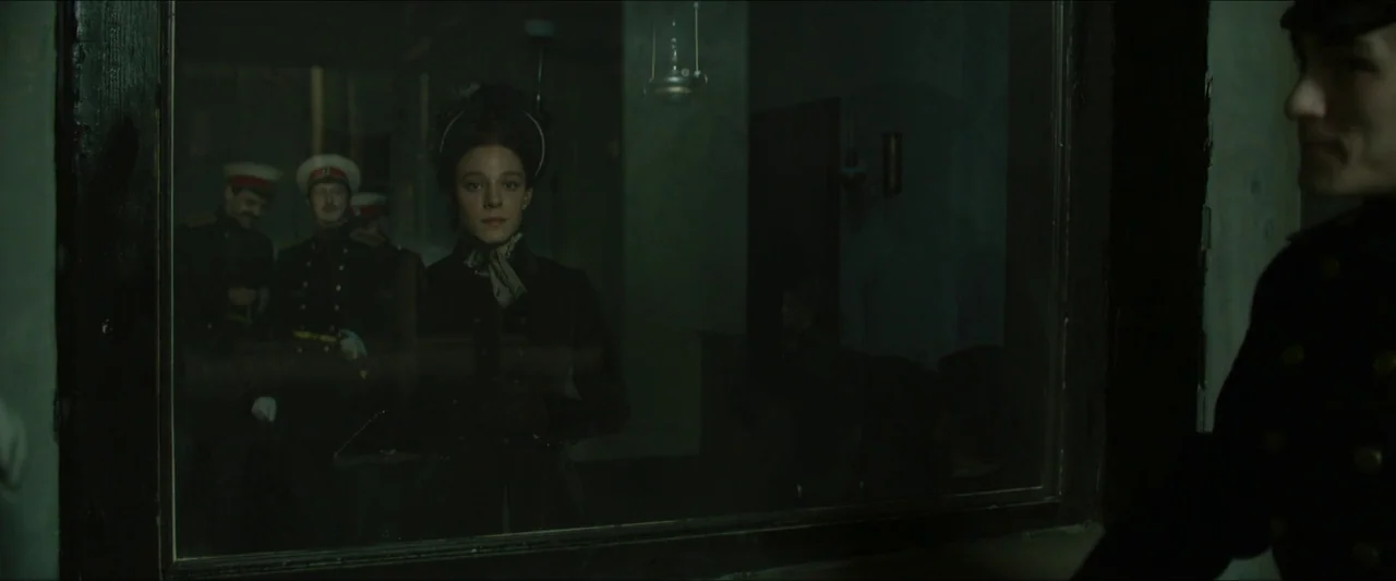 Tchaikovsky’s Wife (2022) movie still: medium — A dimly lit interior space with figures visible behind a large glass partition. A man in uniform…