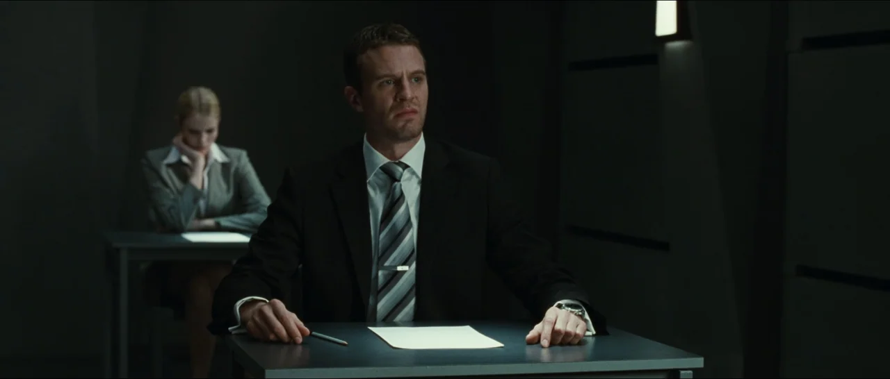 Slackers (2002) movie still: medium — A dimly lit professional setting with two people seated at separate dark desks. A man in the foreground looks…