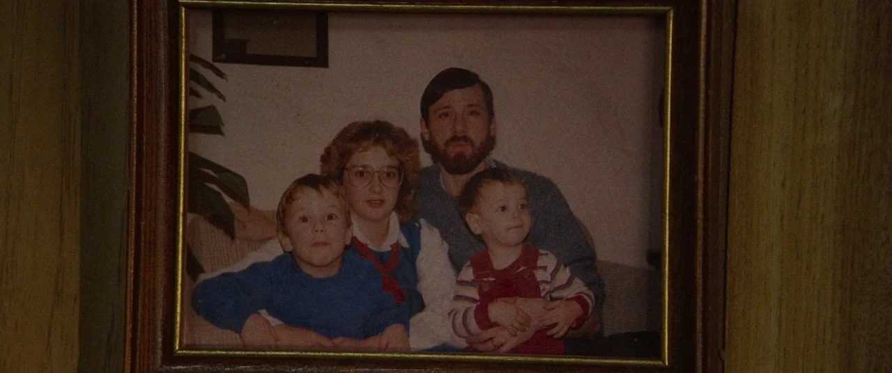 Summer of 84 (2018) movie still: closeup — A framed 1980s photograph showing a man, a woman, and two young boys. The subjects are posing together…